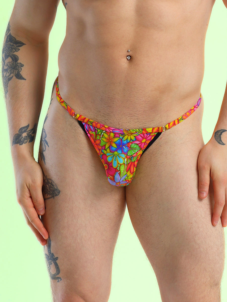 Closeup view of a man wearing a rainbow floral print thong, against a light green background.