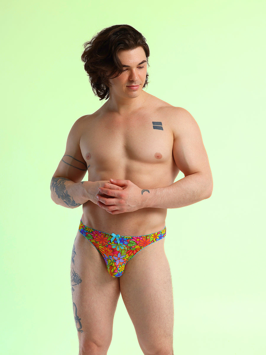 Front view of a man wearing a floral print thong panty with a blue bow, against a green background.