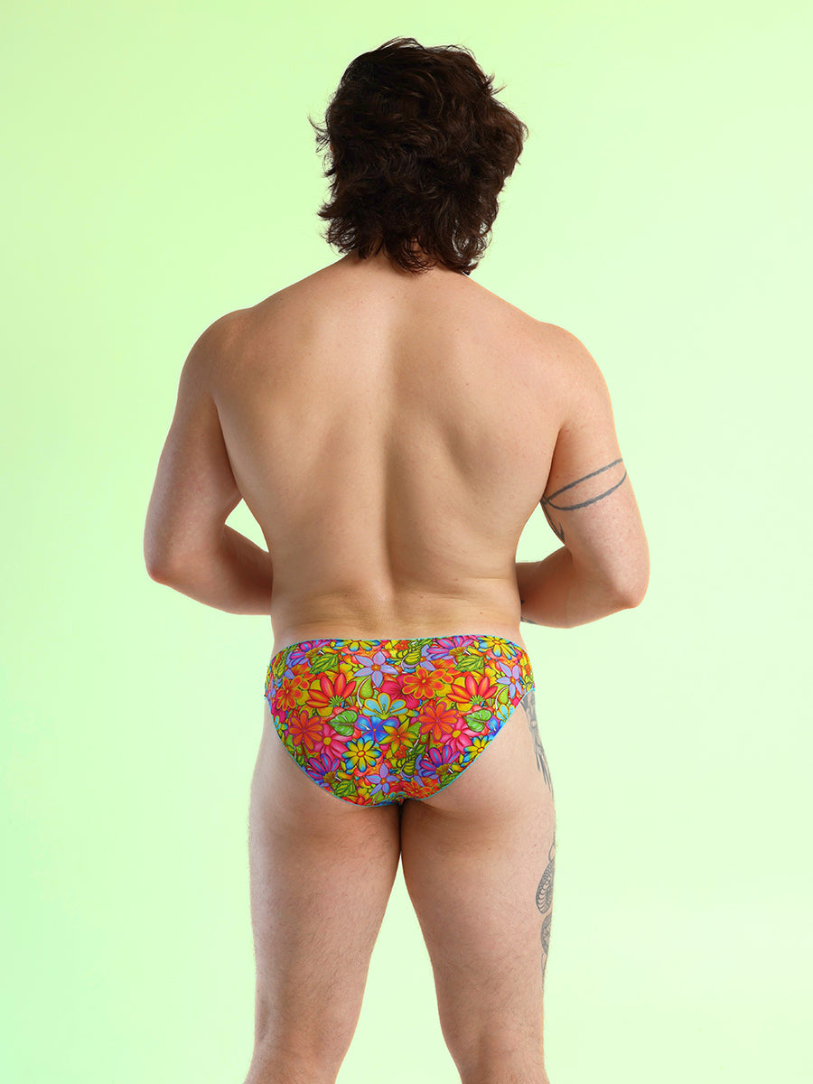 Back view of a man wearing rainbow floral pattern panties with blue lace trim, against a green background.