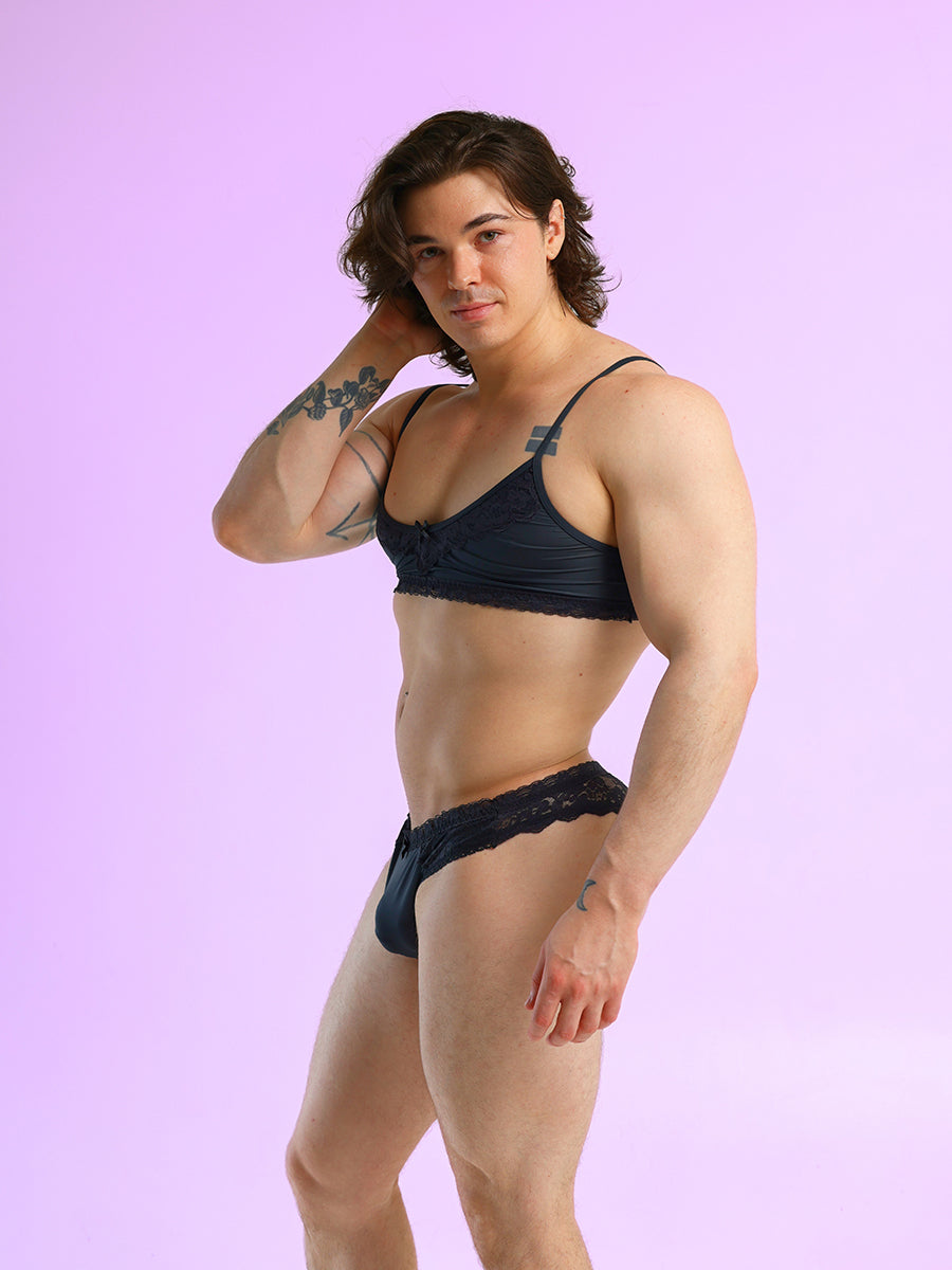 Side view of a man wearing a black bra and matching panties, against a purple background.