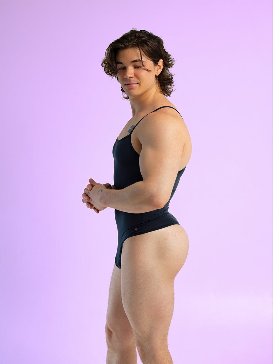 Side view of a man wearing a black thong bodysuit with thin shoulder straps, against a purple background.