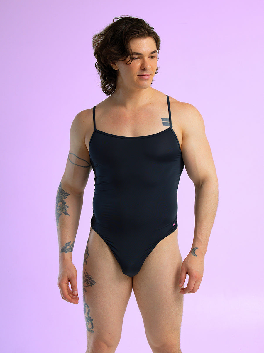 Front view of a man wearing a black thong bodysuit with thin shoulder straps, against a purple background.