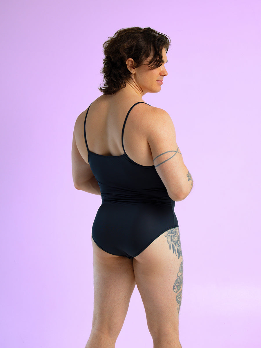 Back view of a man wearing a black bodysuit with thin shoulder straps, against a purple background.