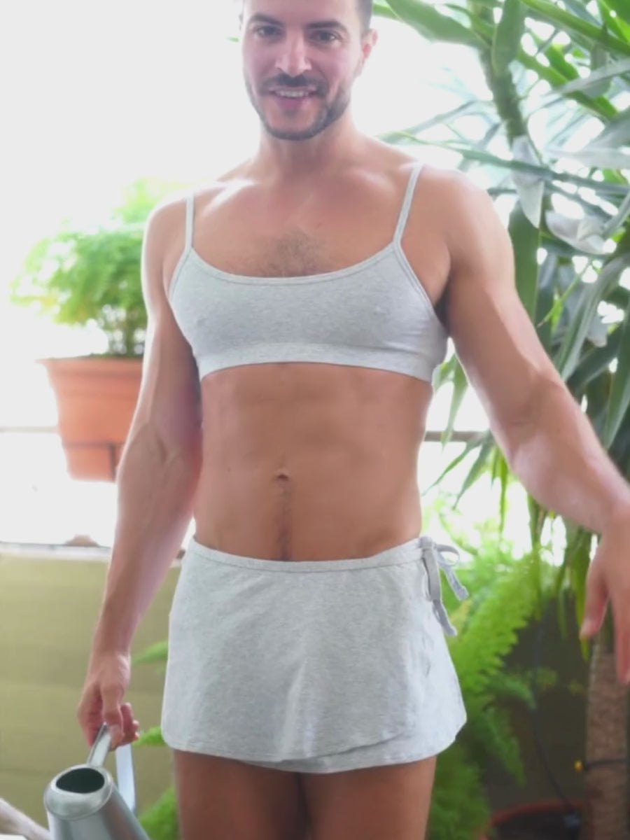 Video of a man wearing a matching grey sports bra and skort, posing outdoors with plants in the background.