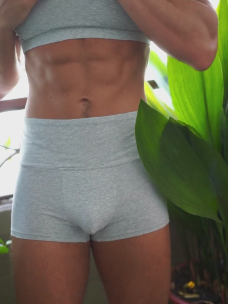 A man wearing matching grey athletic shorts and sports bra, posing outdoors.